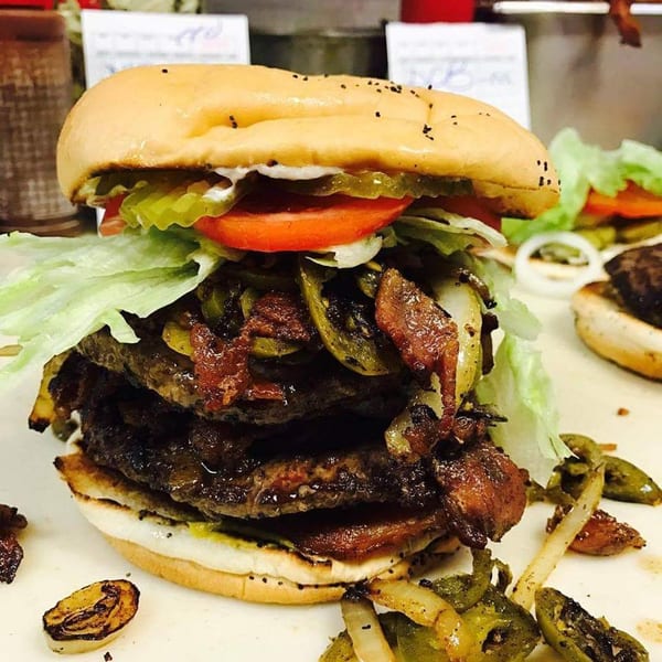 A towering triple-patty burger from Miller's Cafe, loaded with lettuce, tomato, pickles, and grilled onions.