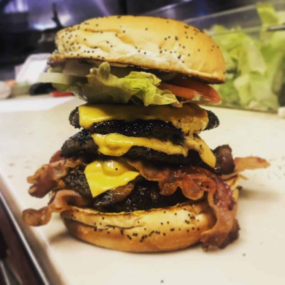 A towering Miller's Cafe triple cheeseburger with bacon, lettuce, tomato, and onion on a poppy seed bun.