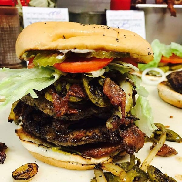 A towering Miller's Cafe burger with multiple patties, bacon, grilled onions, peppers, and fresh toppings.